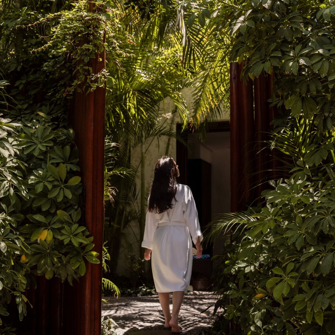 A woman in a white robe walking barefoot through a garden path surrounded by tropical plants.